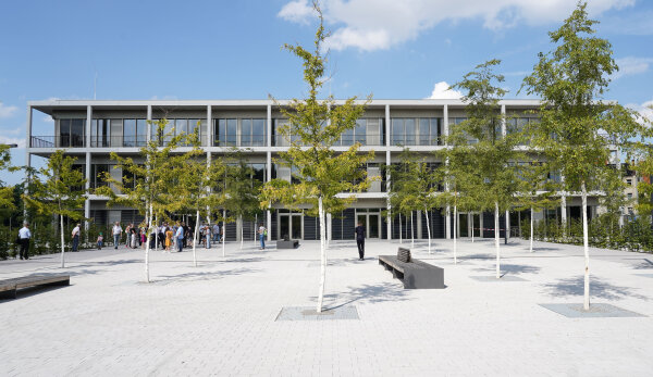 Das Bild zeigt den Vorplatz der neuen Oberschule „Am Hartmannplatz“ mit Sitzbänken und Bäumen. Im Hintergrund ist das Schulgebäude mit Personen zu erkennen.