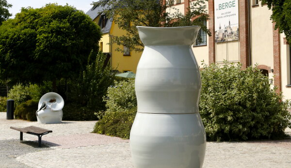 Eine Aufnahme eines Kunstwerkes der Berliner Künstlerin Uli Aigner. Ein vasenförmiges, abstraktes Gebilde aus weißem Porzellan steht auf einem gepflasterten Gehweg. Daneben befinden sich Sitzbänke und Rasen. Im Hintergrund sind Bäume, Büsche und Gebäude erkennbar. Die Sonne scheint.