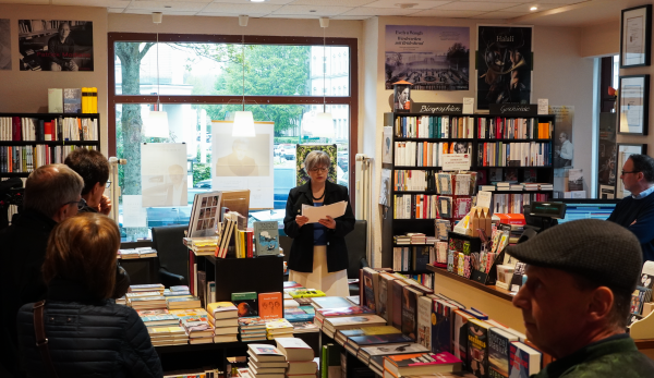 In einem Buchladen steht mittig im Bild eine Frau und liest aus einem Buch vor. Um sie herum stehen Personen und hören zu. Der Raum ist voller Bücherregale und Auslagen mit Büchern. Im Hintergrund befindet sich ein Schaufenster.