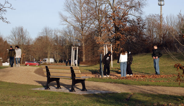 Das Bild zeigt den Bürgerpark Gablenz mit einer grünen Wiese, einem Weg, einer Sitzbank und einem Sportgerät aus schwarzen Stangen mit einer kleinen Gruppe Jugendlicher. Links laufen zwei Personen auf dem Weg. Im Hintergrund sind unbelaubte Bäume erkennbar. Die Sonne scheint.