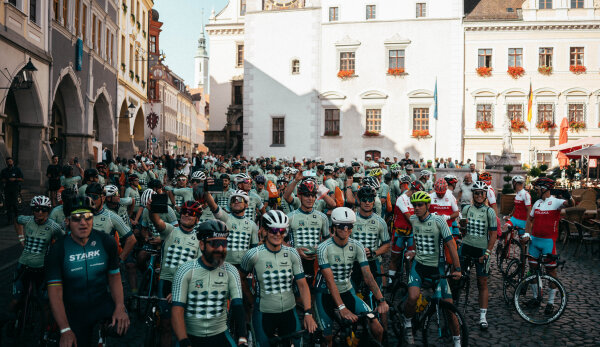 Eine große Gruppe Teilnehmende des European Peace Rides in Fahrradtrikots und -helmen steht mit ihren Fahrrädern in der Innenstadt von Görlitz. Im Hintergrund sind Gebäude der Innenstadt erkennbar. Der Himmel ist blau und wolkenlos.